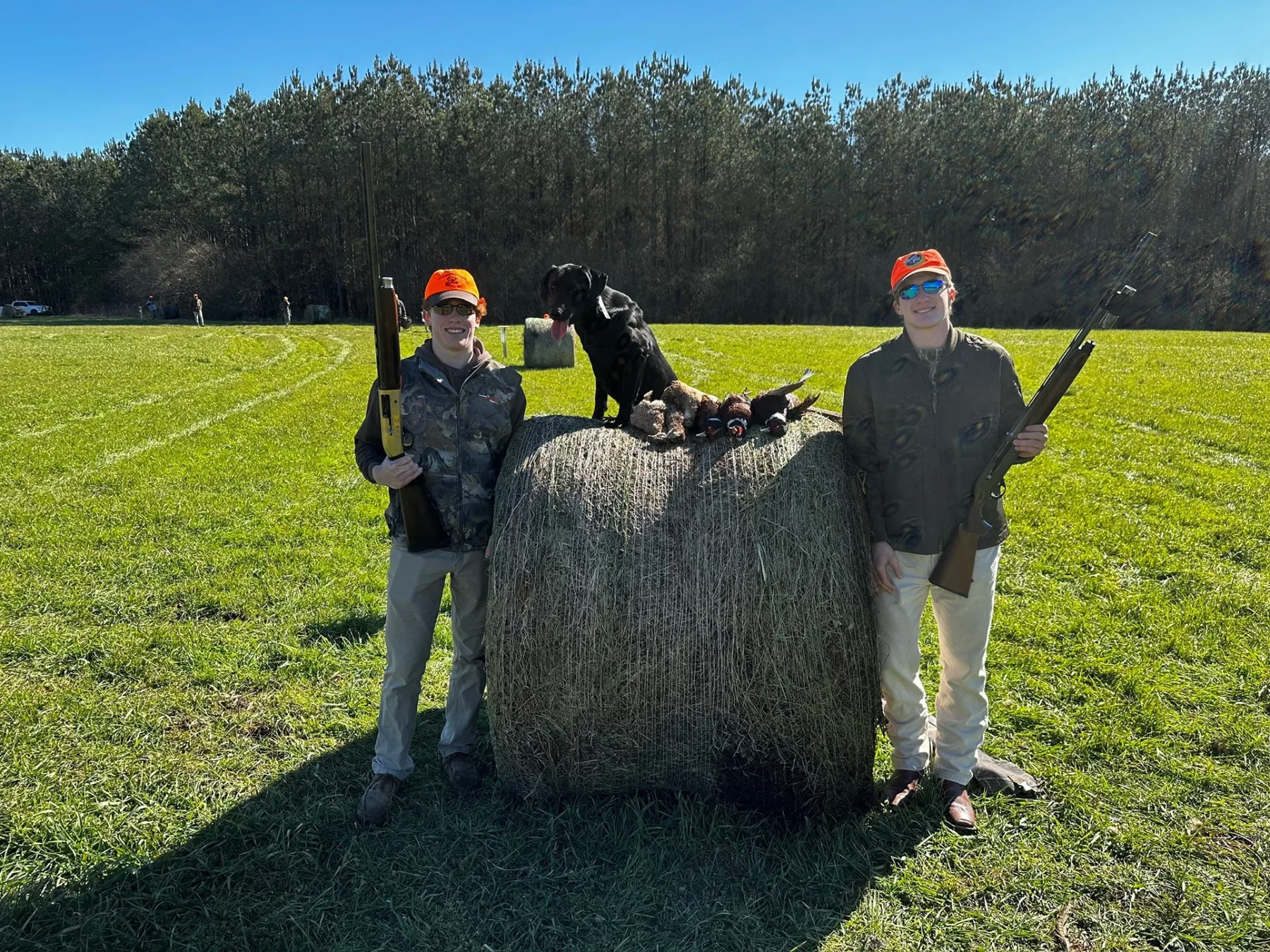 Two shooters and a black Labrador beside a hay bale with harvested pheasants